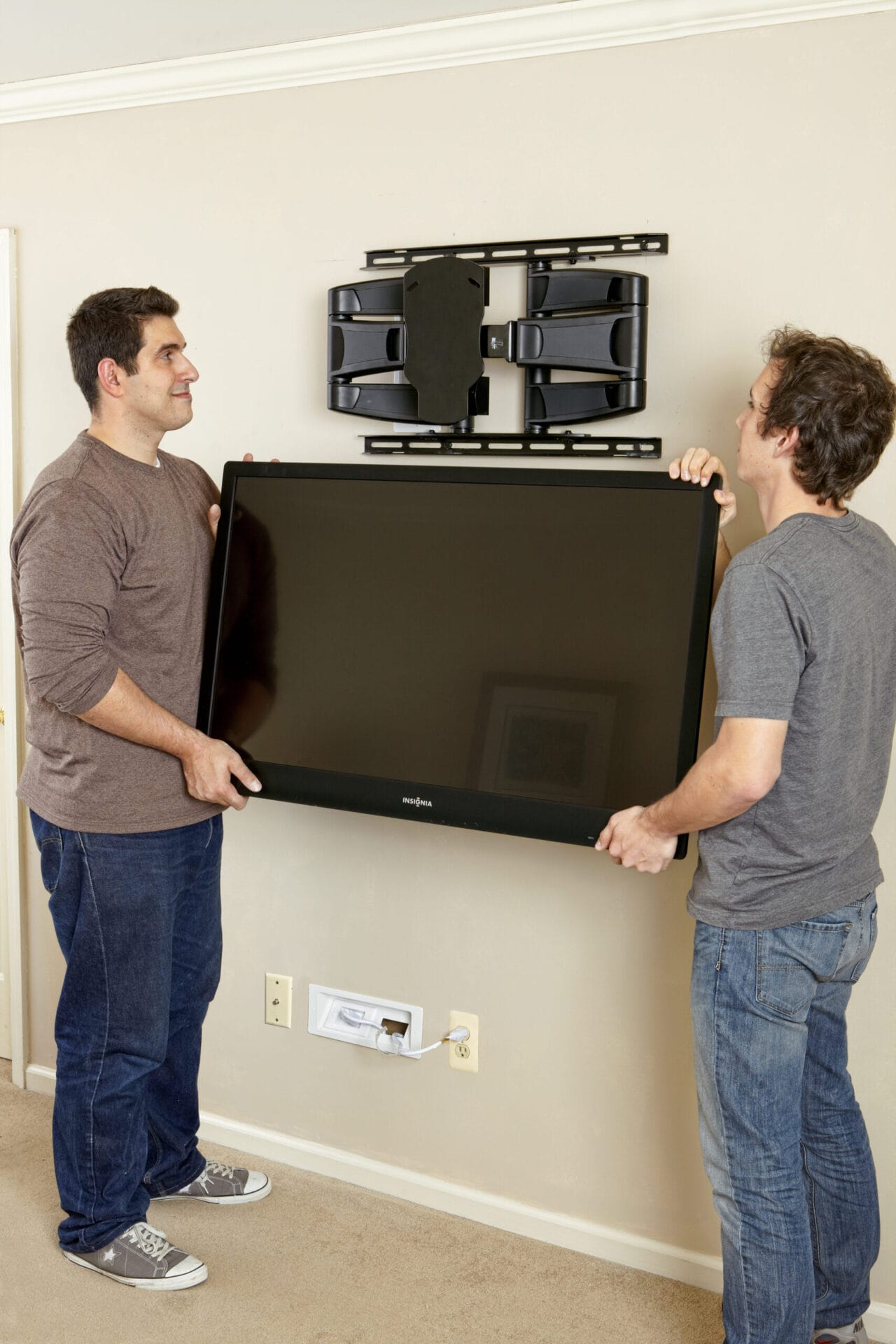 A TV being lifted onto a full-motion mount above a recessed wall outlet.